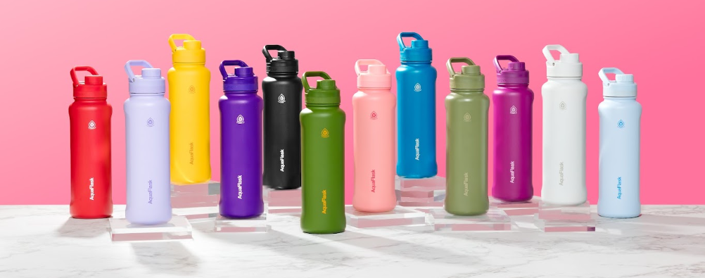 Farverige Aquaflask-vandflasker står på række mod lyserød baggrund, med forskellige låg og håndtag.